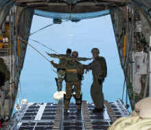 Uniformed people stand on the edge of an aircraft door ready to jump out over water. One person has already started to jump.  